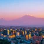 Sunset-lit panorama of Yerevan’s skyline with modern apartment blocks in the foreground and the twin peaks of Mount Ararat glowing atop the horizon.