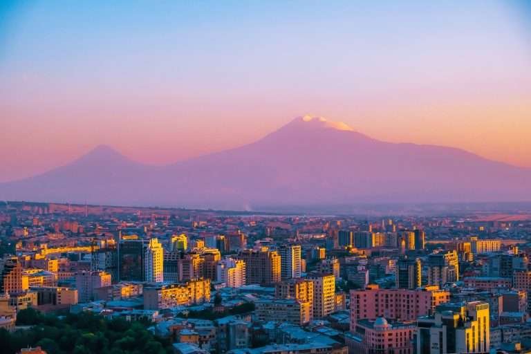 Sunset-lit panorama of Yerevan’s skyline with modern apartment blocks in the foreground and the twin peaks of Mount Ararat glowing atop the horizon.
