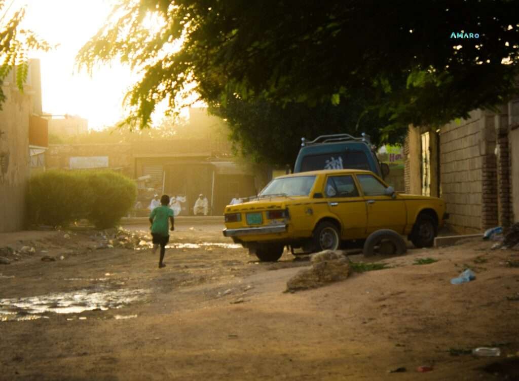 Child running past a broken yellow taxi on an unpaved street in Khartoum, Sudan