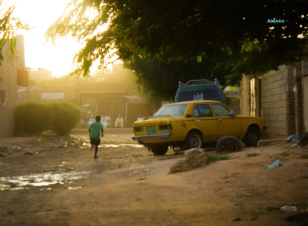 Child running past a broken yellow taxi on an unpaved street in Khartoum, Sudan