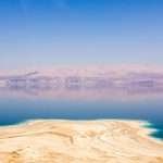 Panoramic view of the Dead Sea with salt deposits in the foreground and mountains in the distance under a clear blue sky.