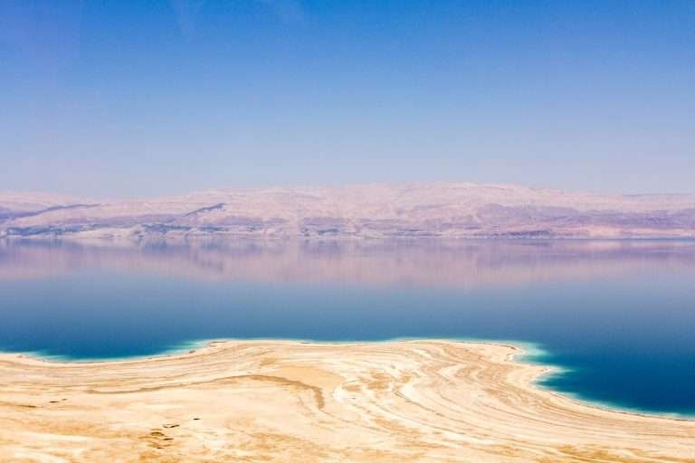 Panoramic view of the Dead Sea with salt deposits in the foreground and mountains in the distance under a clear blue sky.