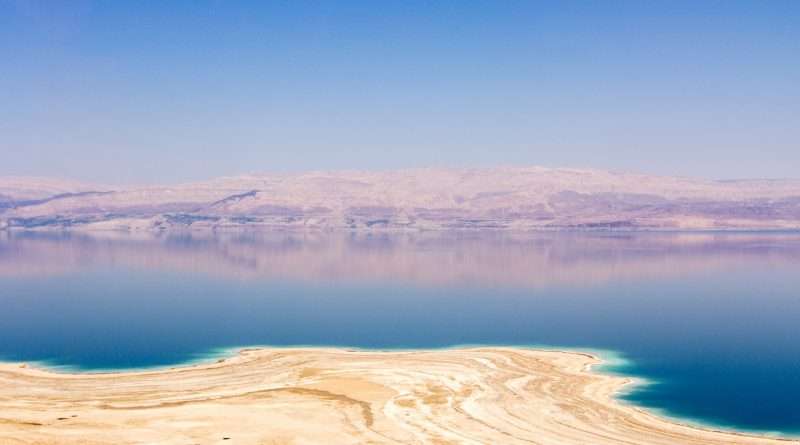 Panoramic view of the Dead Sea with salt deposits in the foreground and mountains in the distance under a clear blue sky.
