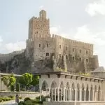 Rabati Castle in Akhaltsikhe, Georgia—stone citadel with crenellated walls and a tall keep overlooking landscaped gardens and a white arched colonnade on a sunny day.