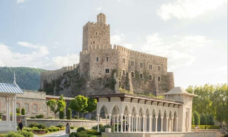 Rabati Castle in Akhaltsikhe, Georgia—stone citadel with crenellated walls and a tall keep overlooking landscaped gardens and a white arched colonnade on a sunny day.