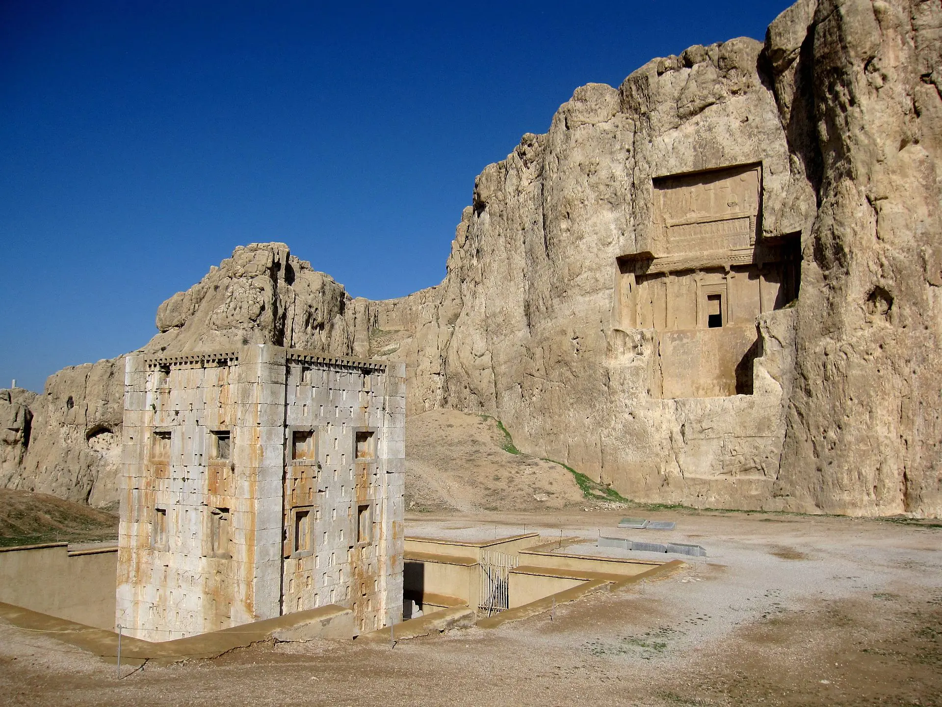Naqsh-e Rustam necropolis showing the white Kaaba-ye Zartosht tower in the foreground and an Achaemenid cliff-tomb façade carved into the limestone cliff at Fars Province, Iran.