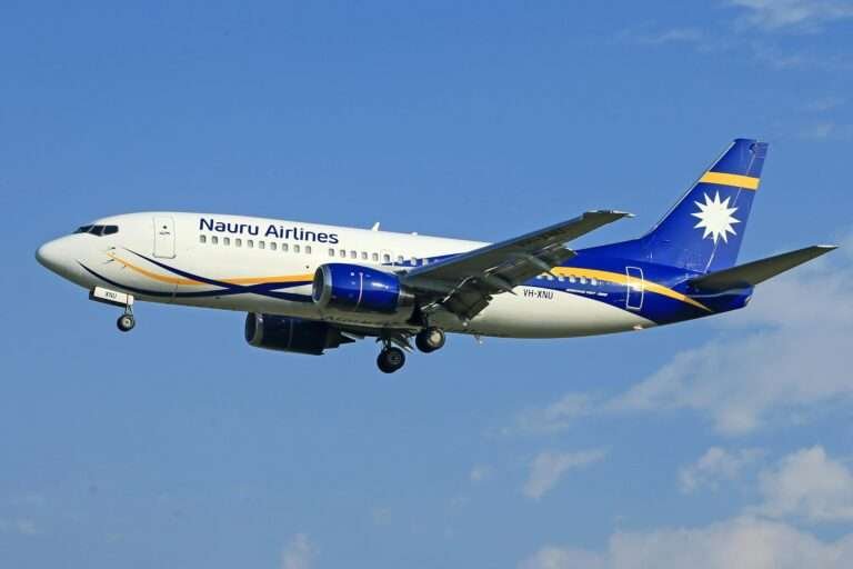 Nauru Airlines Boeing 737-300 aircraft in white, blue, and yellow livery descending against a clear blue sky.