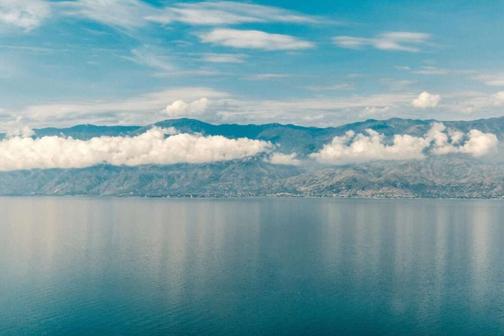Wide-angle view of Lake Tanganyika’s deep blue waters reflecting a ribbon of white clouds, with the rugged mountains of the East African Rift rising along the distant shoreline under a partly cloudy sky.