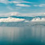Wide-angle view of Lake Tanganyika’s deep blue waters reflecting a ribbon of white clouds, with the rugged mountains of the East African Rift rising along the distant shoreline under a partly cloudy sky.