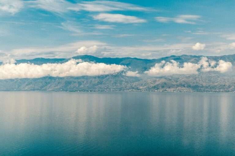 Wide-angle view of Lake Tanganyika’s deep blue waters reflecting a ribbon of white clouds, with the rugged mountains of the East African Rift rising along the distant shoreline under a partly cloudy sky.