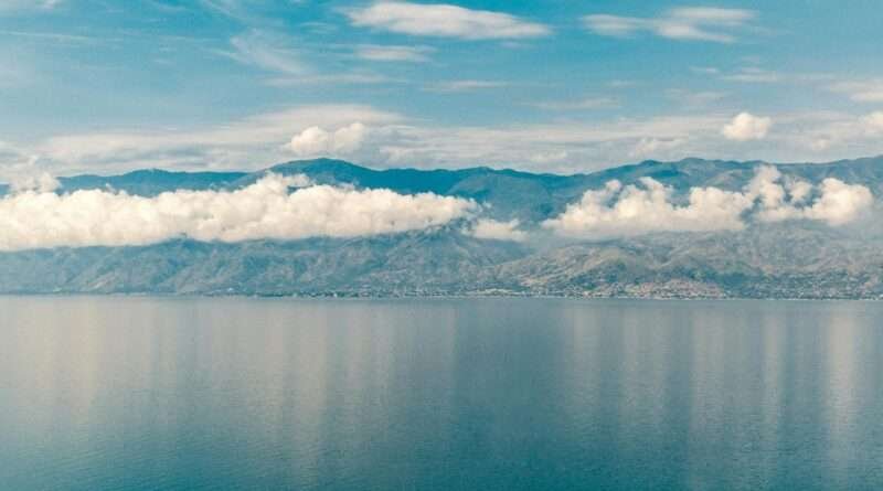 Wide-angle view of Lake Tanganyika’s deep blue waters reflecting a ribbon of white clouds, with the rugged mountains of the East African Rift rising along the distant shoreline under a partly cloudy sky.