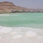 Salt formations along the Dead Sea shoreline with turquoise water and desert mountains in the background.