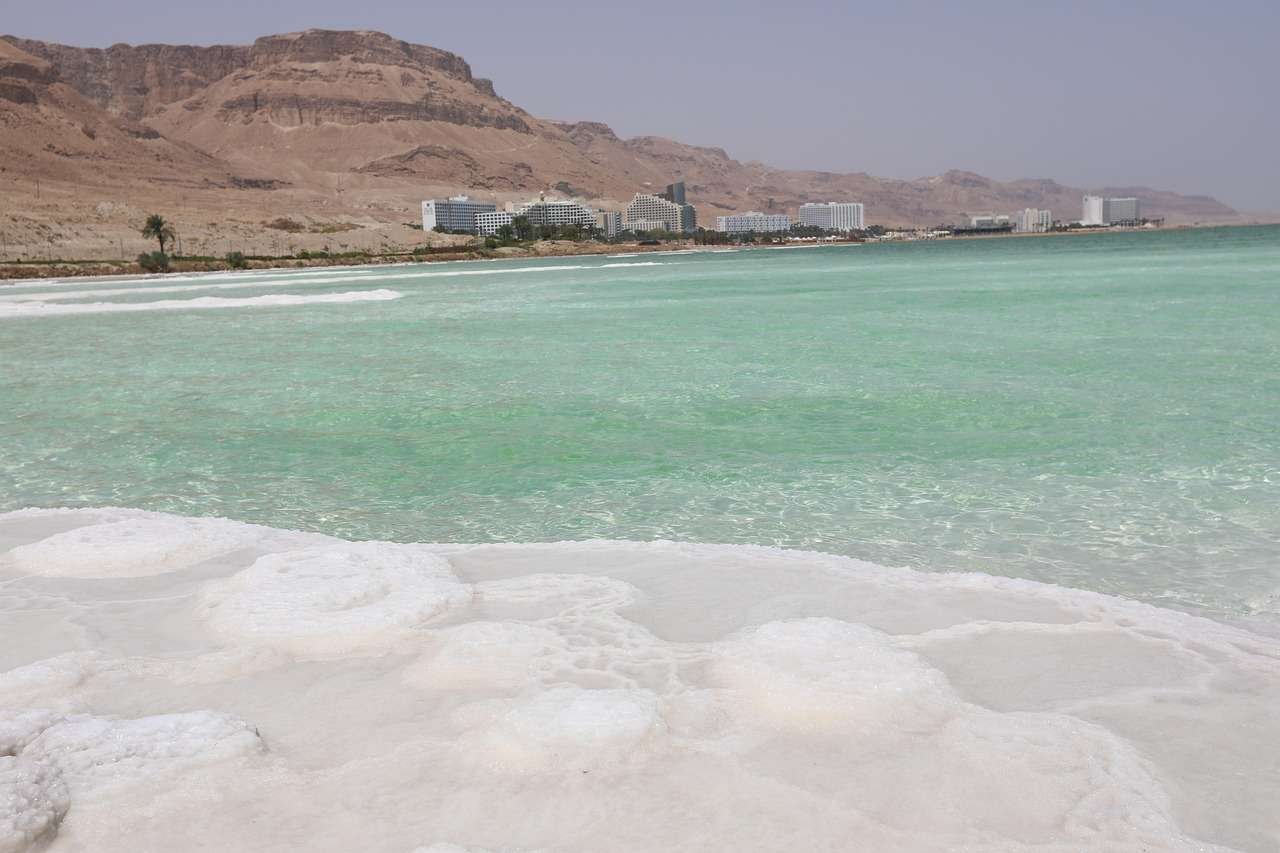 Salt formations along the Dead Sea shoreline with turquoise water and desert mountains in the background.