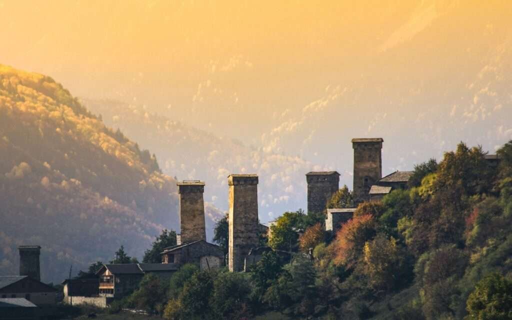 Svan defensive towers above Mestia, Upper Svaneti, Georgia, at golden-hour sunset with autumn forests and layered Caucasus mountains behind.
