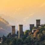 Svan defensive towers above Mestia, Upper Svaneti, Georgia, at golden-hour sunset with autumn forests and layered Caucasus mountains behind.