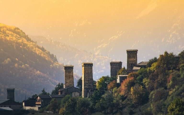 Svan defensive towers above Mestia, Upper Svaneti, Georgia, at golden-hour sunset with autumn forests and layered Caucasus mountains behind.