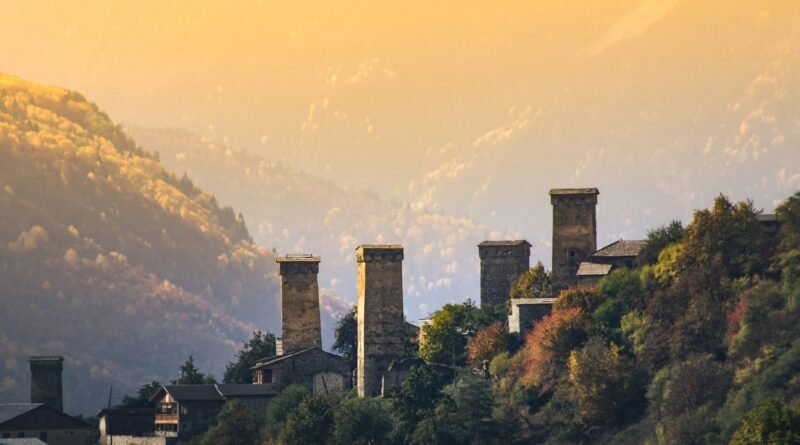 Svan defensive towers above Mestia, Upper Svaneti, Georgia, at golden-hour sunset with autumn forests and layered Caucasus mountains behind.