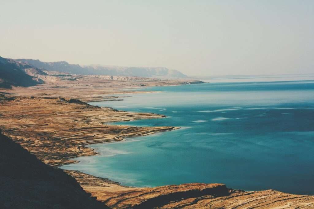 A wide-angle view of the Dead Sea shoreline in Israel, showing turquoise waters, layered salt terraces, and arid desert mountains under a hazy sky.