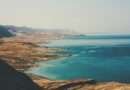 A wide-angle view of the Dead Sea shoreline in Israel, showing turquoise waters, layered salt terraces, and arid desert mountains under a hazy sky.
