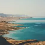 A wide-angle view of the Dead Sea shoreline in Israel, showing turquoise waters, layered salt terraces, and arid desert mountains under a hazy sky.