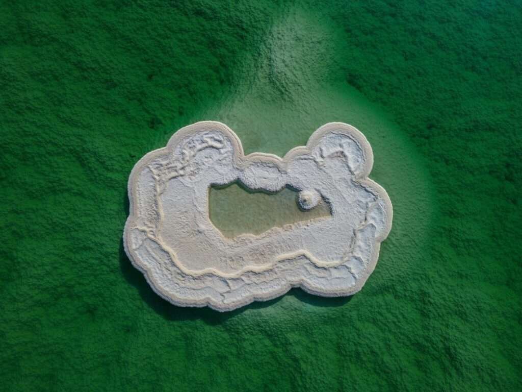 Aerial view of a white salt island formation in the green waters of the Dead Sea, showing crystallized magnesium and mineral deposits.