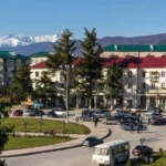 Tskhinvali cityscape with urban buildings, traffic, evergreen trees and snow-covered Greater Caucasus mountains.