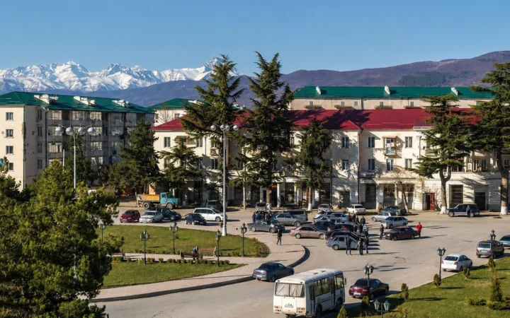 Tskhinvali cityscape with urban buildings, traffic, evergreen trees and snow-covered Greater Caucasus mountains.
