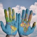 World map painted on two open hands against a cloudy sky, symbolizing people–environment relationships at the heart of geography.