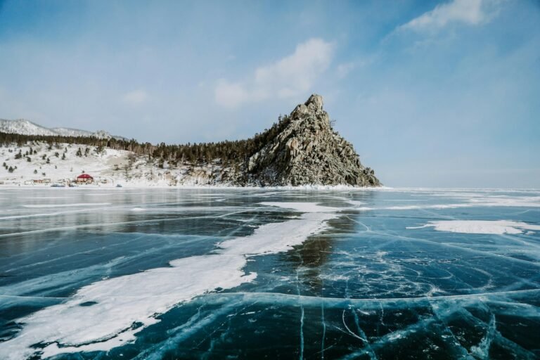 Rocky headland and forested shore rising above the blue, cracked ice of frozen Lake Baikal under a bright winter sky.