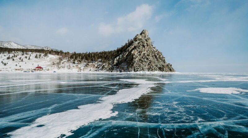 Rocky headland and forested shore rising above the blue, cracked ice of frozen Lake Baikal under a bright winter sky.