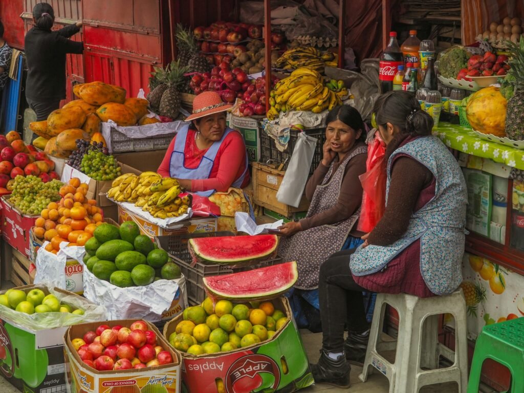Three women chatting at a neighborhood fruit stall in La Paz, Bolivia, surrounded by crates of bananas, avocados, papayas, apples, and watermelon.