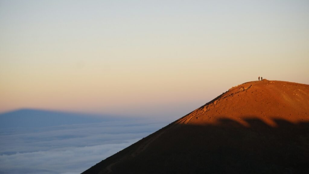 Sunset hikers on a Mauna Kea cinder cone above a sea of clouds, long mountain shadow on the slope near the ~13,803-ft (4,207-m) summit
