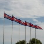 Row of North Korean (DPRK) flags flying on tall poles against a cloudy sky.