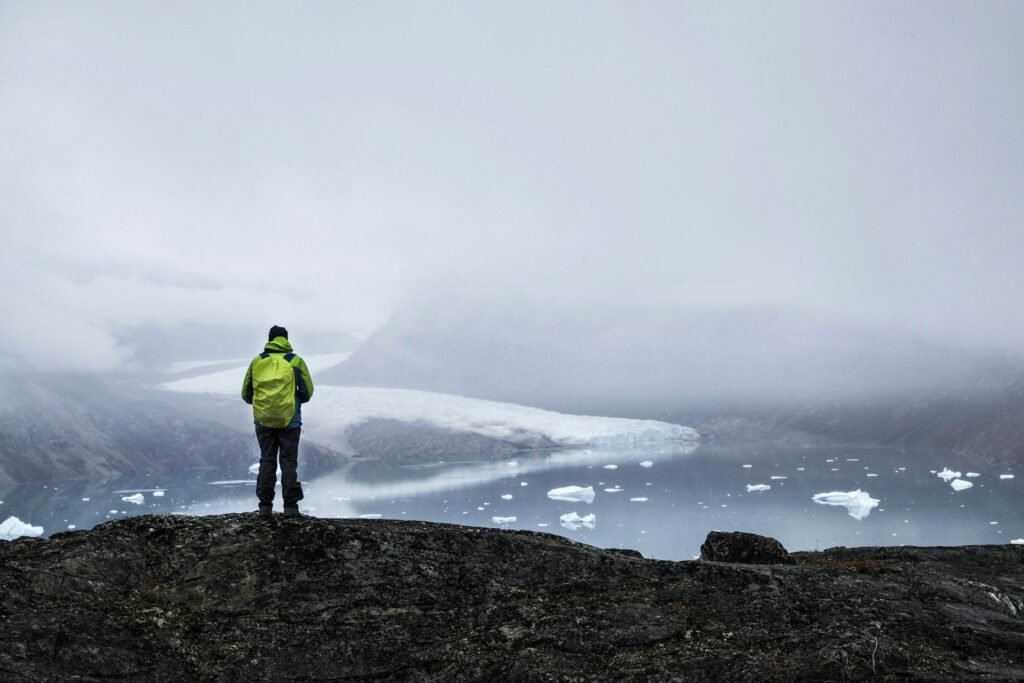 Person overlooking a foggy Greenland fjord with a tidewater glacier and scattered ice Title: Greenland’s Hidden Geography — Glacier and Fjord Vista