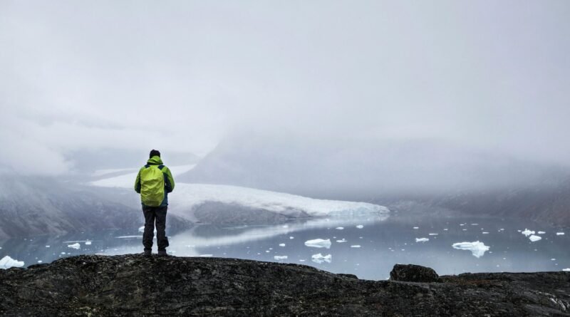 Person overlooking a foggy Greenland fjord with a tidewater glacier and scattered ice Title: Greenland’s Hidden Geography — Glacier and Fjord Vista