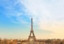 Eiffel Tower centered from the Trocadéro at sunset, blue-to-gold sky over Paris with base arches, carousel, and winter trees in view.