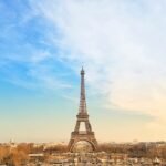 Eiffel Tower centered from the Trocadéro at sunset, blue-to-gold sky over Paris with base arches, carousel, and winter trees in view.