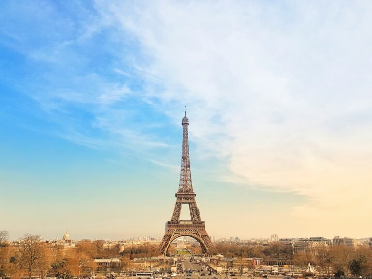 Eiffel Tower centered from the Trocadéro at sunset, blue-to-gold sky over Paris with base arches, carousel, and winter trees in view.