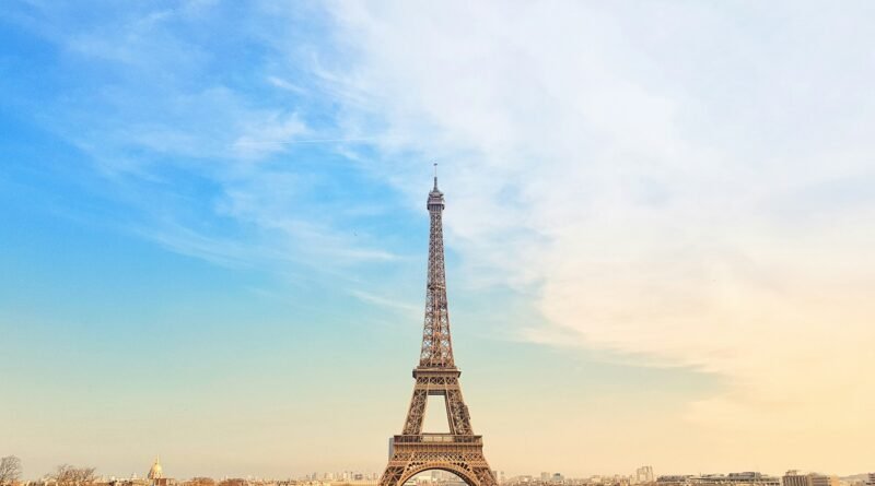Eiffel Tower centered from the Trocadéro at sunset, blue-to-gold sky over Paris with base arches, carousel, and winter trees in view.