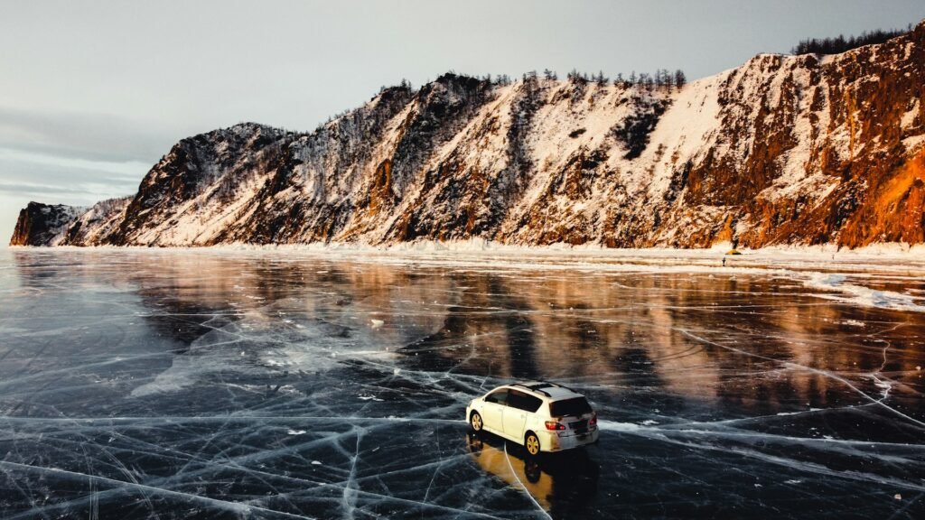 White car driving on the cracked, mirror-like ice of frozen Lake Baikal below steep snow-covered cliffs at sunset.