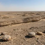 Wind-carved yardang ridges in a desert basin, with rounded stones on a gravelly surface in the foreground.