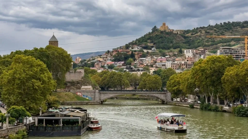 Mtkvari River flowing through Tbilisi, Georgia, with a bridge, riverside trees, and the old city rising on the hillside.