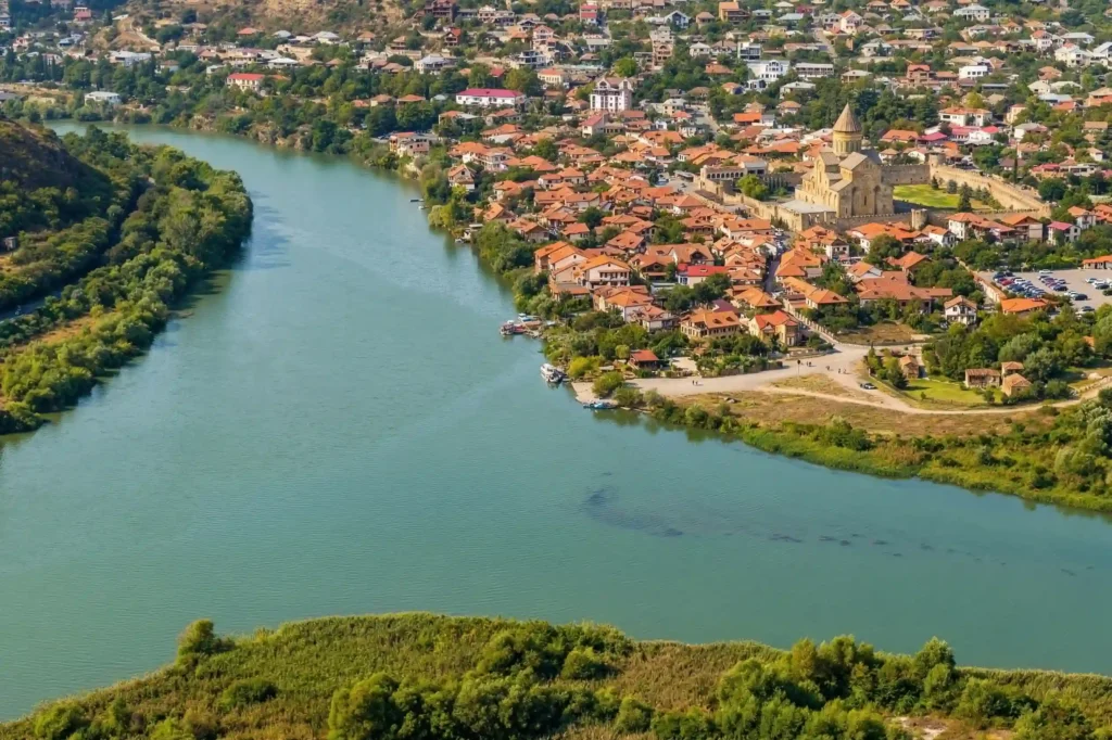 Aerial view of Mtskheta, Georgia, beside the Mtkvari and Aragvi river corridor, with historic buildings and a cathedral near the water.