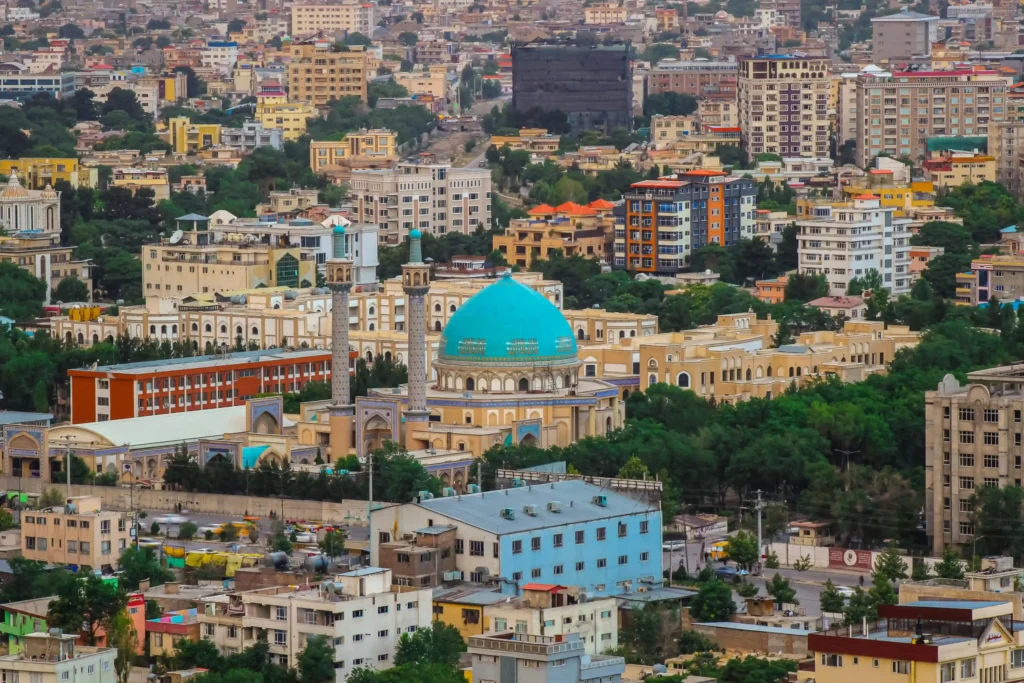 Blue-domed mosque in Kabul with dense urban neighborhoods and city buildings in the background.