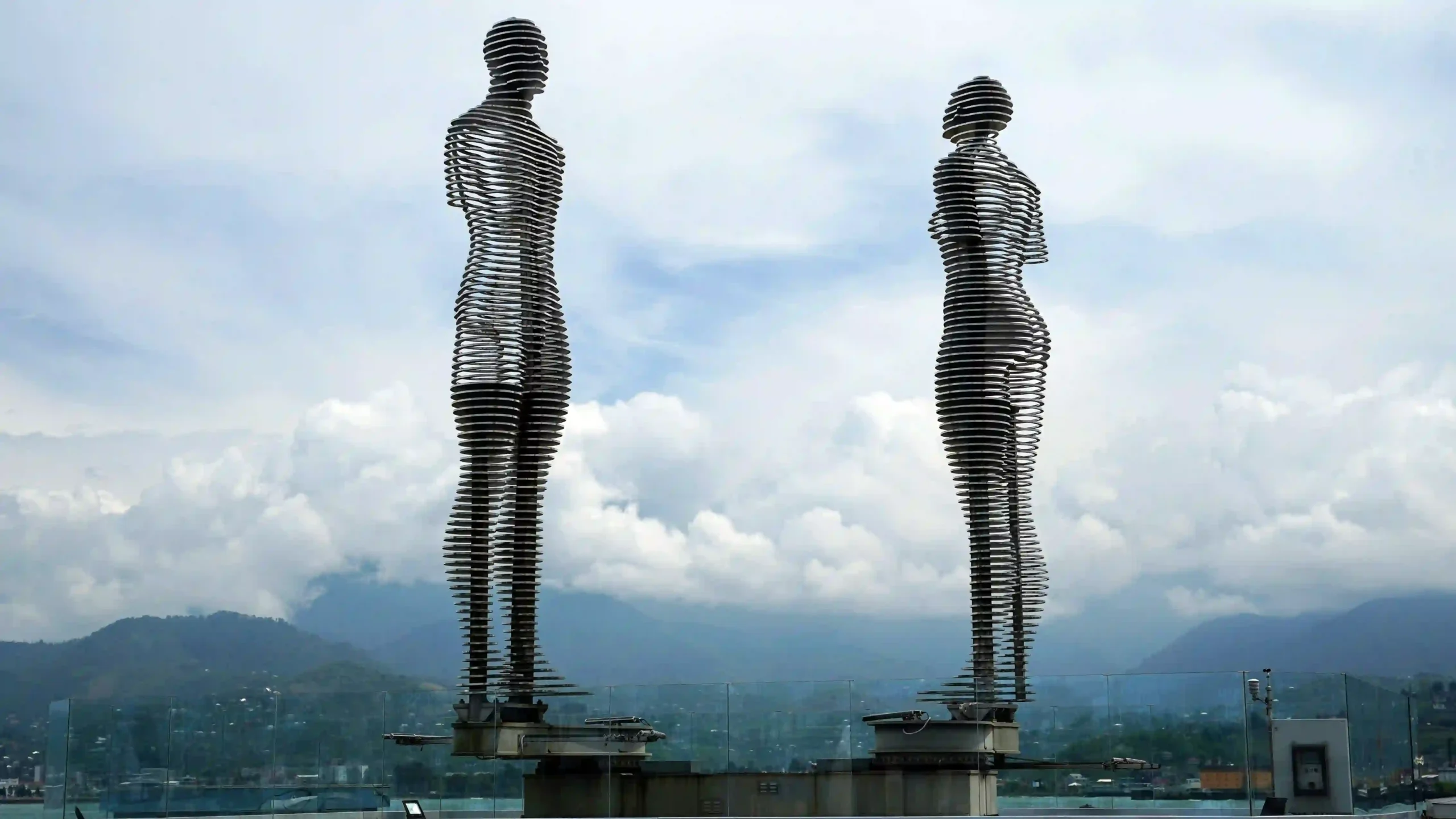 Ali and Nino metal sculptures on Batumi’s waterfront, with cloud-covered mountains behind them.